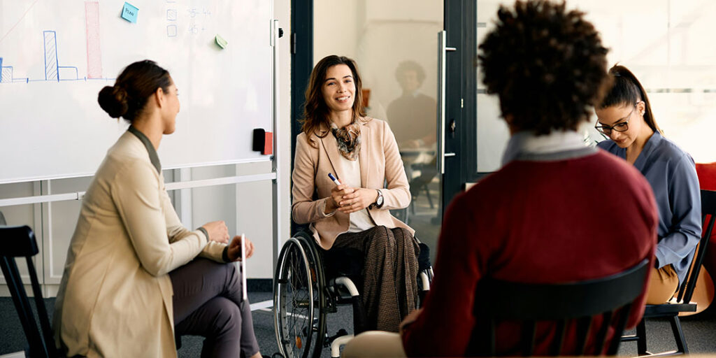 A woman in a wheelchair is in front of a whiteboard holding a dry-erase marker and talking with three people